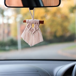 May include: A white macrame car charm with a wooden stick and tassels hanging from the rearview mirror of a car.