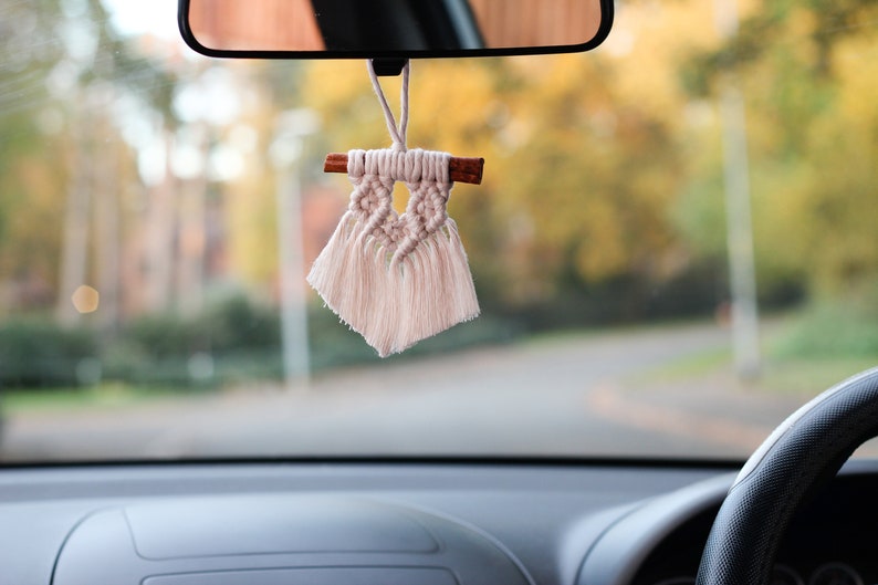 May include: A white macrame car charm with a wooden stick and tassels hanging from the rearview mirror of a car.