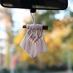 May include: A white macrame car air freshener with a cinnamon stick hanging from a rearview mirror. The background is blurred and shows trees in the fall.