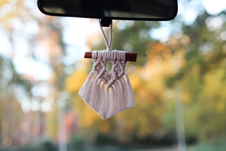 May include: A white macrame car air freshener with a cinnamon stick hanging from a rearview mirror. The background is blurred and shows trees in the fall.