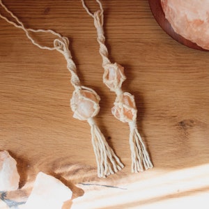 May include: Two pink Himalayan salt crystals are suspended in macrame string with white tassels. The crystals are hanging from a wooden surface.