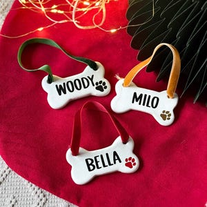 Puede incluir: Tres adornos blancos de cerámica con forma de hueso para perros con los nombres "WOODY", "MILO" y "BELLA" en fuente negra. Cada hueso tiene una huella de pata y una cinta para colgar. El fondo es rojo con una guirnalda de luces.
