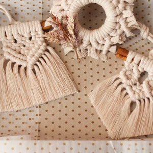 May include: Two handmade macrame ornaments with fringe, each attached to a cinnamon stick. The ornaments are cream colored and are displayed on a white background with gold polka dots.