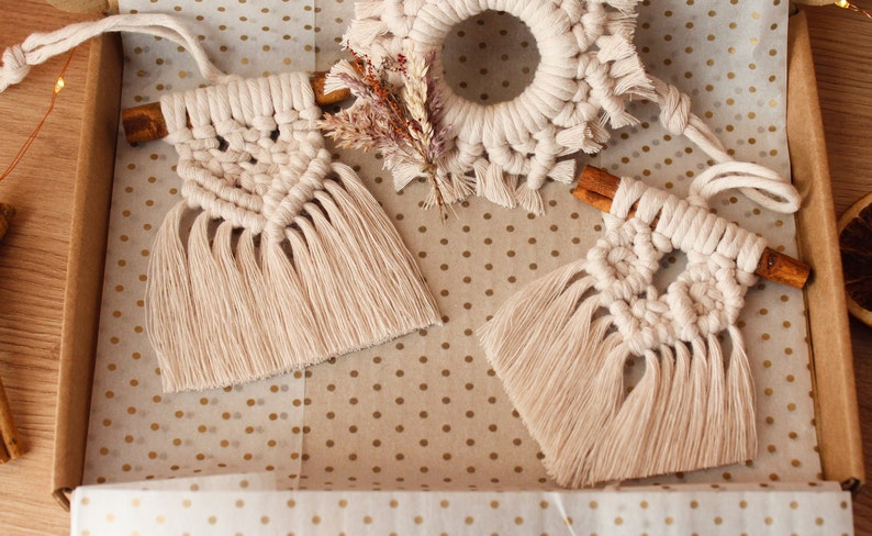 May include: Two handmade macrame ornaments with fringe, each attached to a cinnamon stick. The ornaments are cream colored and are displayed on a white background with gold polka dots.