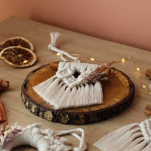 May include: Three macrame ornaments with fringe, one on a wooden slice, one with dried flowers, and one with a cinnamon stick. The ornaments are white and orange.