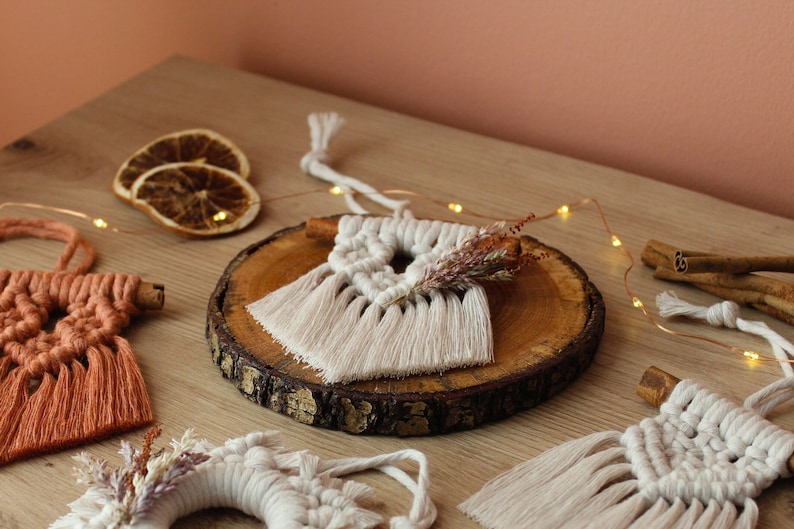 May include: Three macrame ornaments with fringe, one on a wooden slice, one with dried flowers, and one with a cinnamon stick. The ornaments are white and orange.
