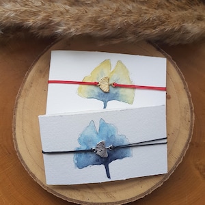 Könnte beinhalten: Zwei handgefertigte Armbänder mit Ginkgoblatt-Anhängern. Eines hat ein rotes Band und einen goldfarbenen Anhänger, das andere ein schwarzes Band und einen silberfarbenen Anhänger. Sie werden auf weißen Karten mit Aquarellmalereien von Ginkgoblättern präsentiert.