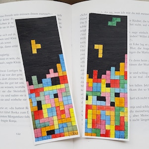 May include: Two watercolour paintings of a classic video game, Tetris, on a book. The game pieces are in various colours, including red, yellow, blue, green, pink, and black. The background of the paintings is black.