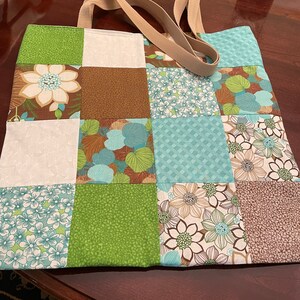 K&ouml;nnte beinhalten: Eine Patchwork-Tragetasche mit braunen, gr&uuml;nen und blauen Blumenmustern. Die Tasche hat zwei beigefarbene Stoffgriffe.