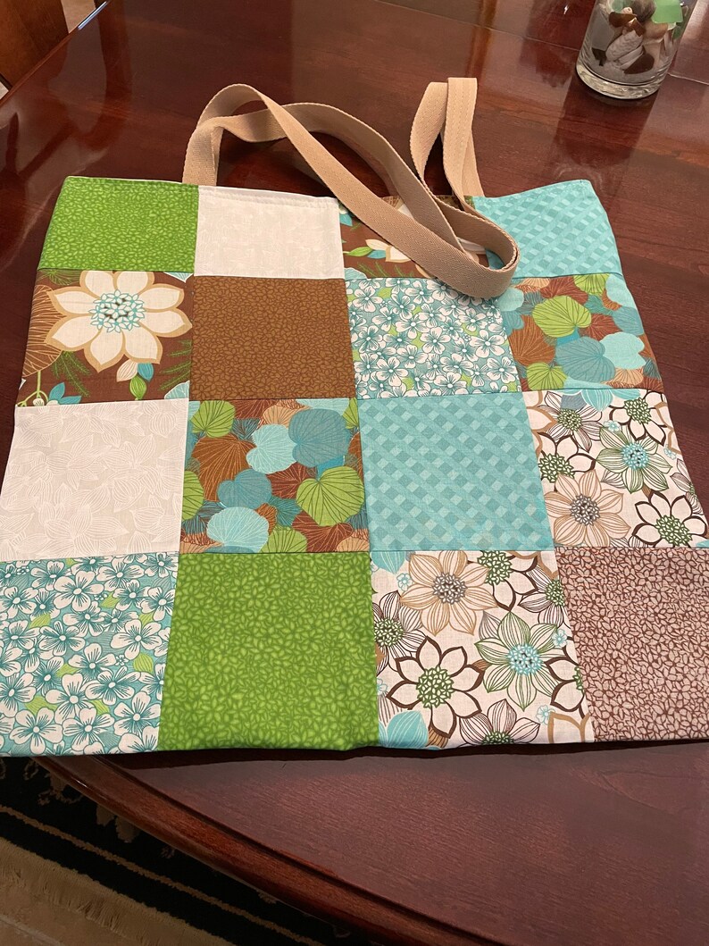 K&ouml;nnte beinhalten: Eine Patchwork-Tragetasche mit braunen, gr&uuml;nen und blauen Blumenmustern. Die Tasche hat zwei beigefarbene Stoffgriffe.