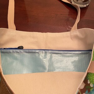K&ouml;nnte beinhalten: Eine beige Canvas-Tragetasche mit einem Rei&szlig;verschlussfach und einem transparenten Plastik-Mesh-Futter. Die Tasche hat ein Blumenmuster auf dem Boden und einen braunen Riemen.