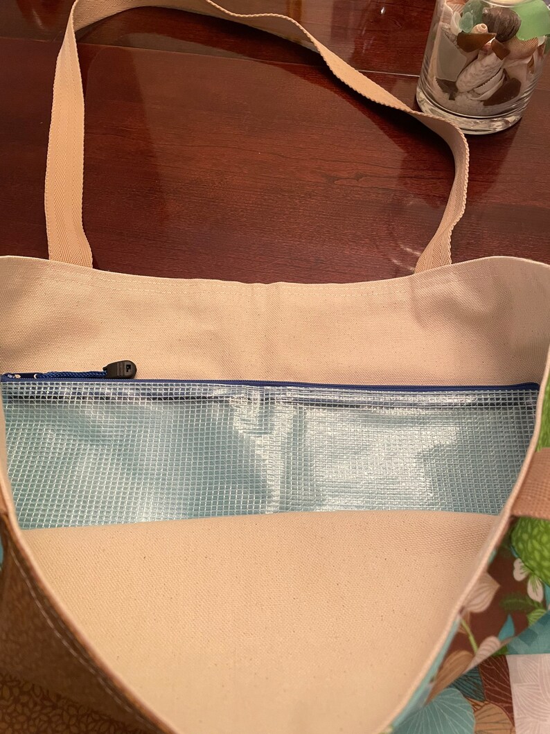 K&ouml;nnte beinhalten: Eine beige Canvas-Tragetasche mit einem Rei&szlig;verschlussfach und einem transparenten Plastik-Mesh-Futter. Die Tasche hat ein Blumenmuster auf dem Boden und einen braunen Riemen.