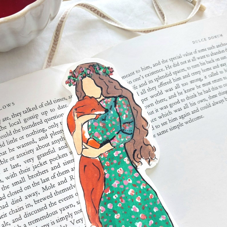 Pu&ograve; includere: Un segnalibro stravagante con un'illustrazione di una donna in un vestito floreale verde che abbraccia una volpe. La donna ha lunghi capelli castani e una corona floreale. Il segnalibro &egrave; posto su un libro con testo.