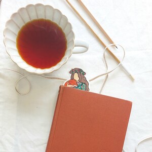 Pu&ograve; includere: Vista dall'alto di un libro arancione con un segnalibro raffigurante una donna che tiene una volpe. Una tazza bianca con bordo smerlato contiene t&egrave; marrone scuro. Sono visibili anche materiali artistici sul tavolo.