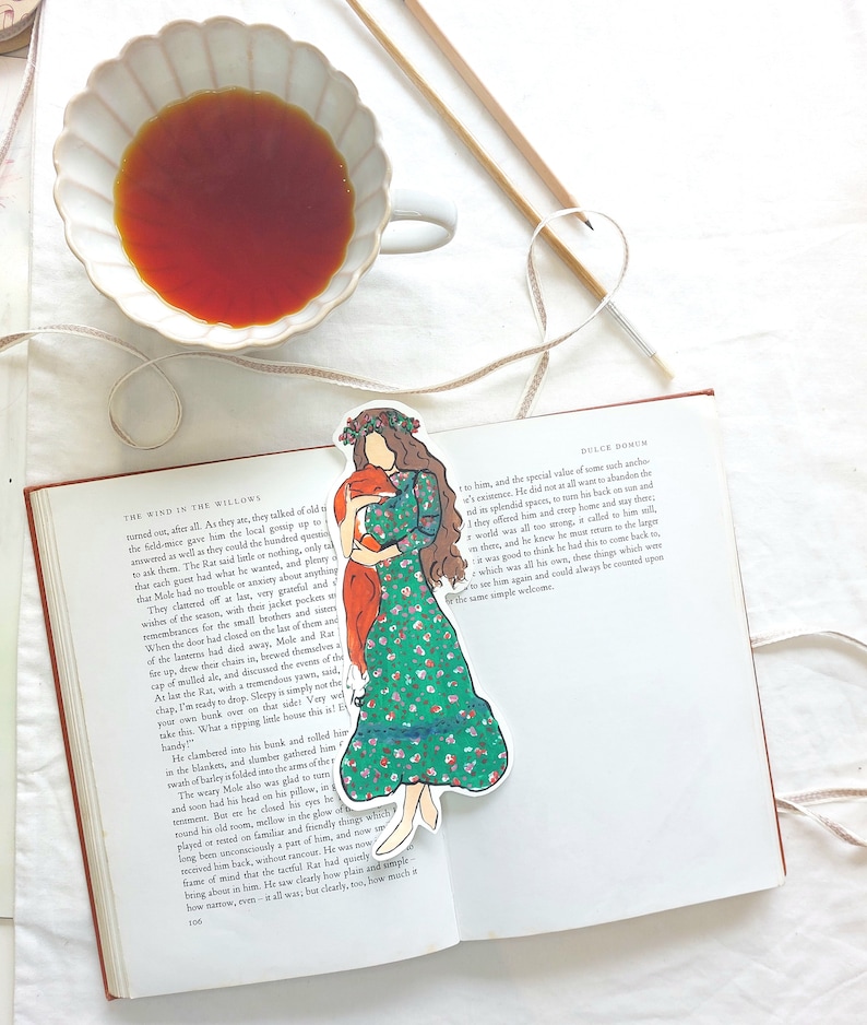 Pu&ograve; includere: Un segnalibro con una donna in un vestito floreale verde che abbraccia una volpe, posto su un libro aperto. Una tazza di t&egrave; e ferri da maglia sono sullo sfondo. Il libro &egrave; intitolato "The Wind in the Willows".