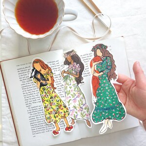Pu&ograve; includere: Tre segnalibri illustrati raffiguranti donne in abiti floreali, che tengono in mano un animale nero, fiori e una sciarpa rossa. I segnalibri sono su un libro aperto, con una tazza di t&egrave; e bacchette sullo sfondo.