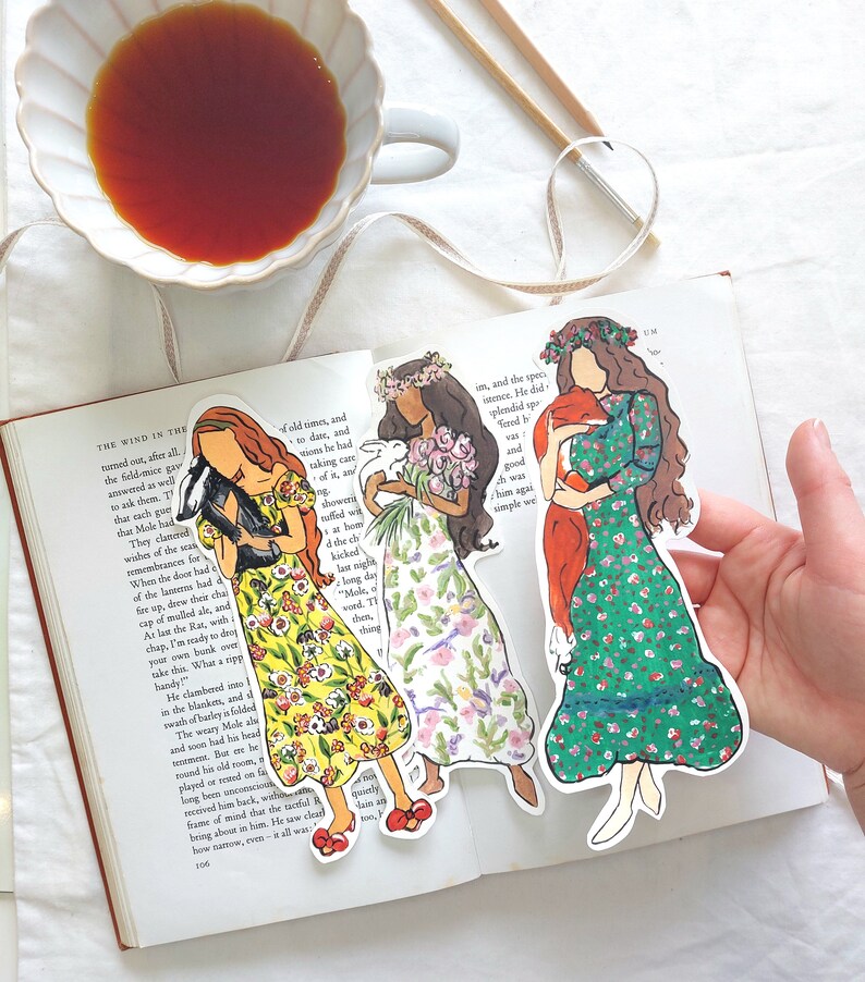 Pu&ograve; includere: Tre segnalibri illustrati raffiguranti donne in abiti floreali, che tengono in mano un animale nero, fiori e una sciarpa rossa. I segnalibri sono su un libro aperto, con una tazza di t&egrave; e bacchette sullo sfondo.