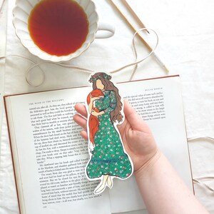 Pu&ograve; includere: Un segnalibro colorato con una donna in un vestito floreale verde che tiene una volpe. Il segnalibro &egrave; tenuto sopra un libro aperto. Una tazza di t&egrave; e delle matite sono sullo sfondo.