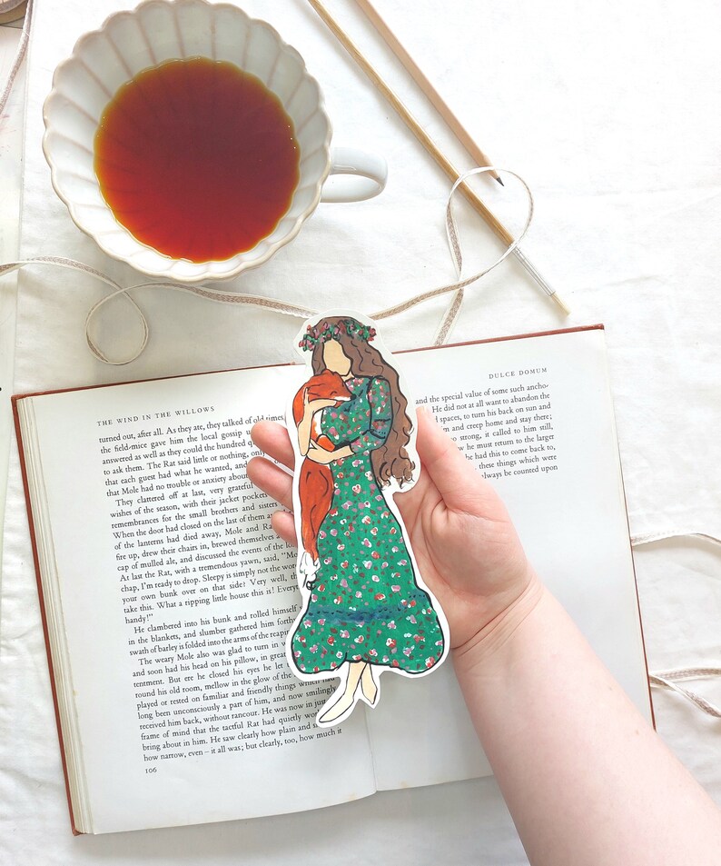 Pu&ograve; includere: Un segnalibro colorato con una donna in un vestito floreale verde che tiene una volpe. Il segnalibro &egrave; tenuto sopra un libro aperto. Una tazza di t&egrave; e delle matite sono sullo sfondo.