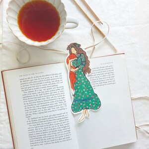 Pu&ograve; includere: Un segnalibro stravagante con una donna in un vestito floreale verde che tiene in braccio una volpe. Il segnalibro &egrave; posto su un libro aperto, insieme a una tazza di t&egrave; e elementi decorativi.
