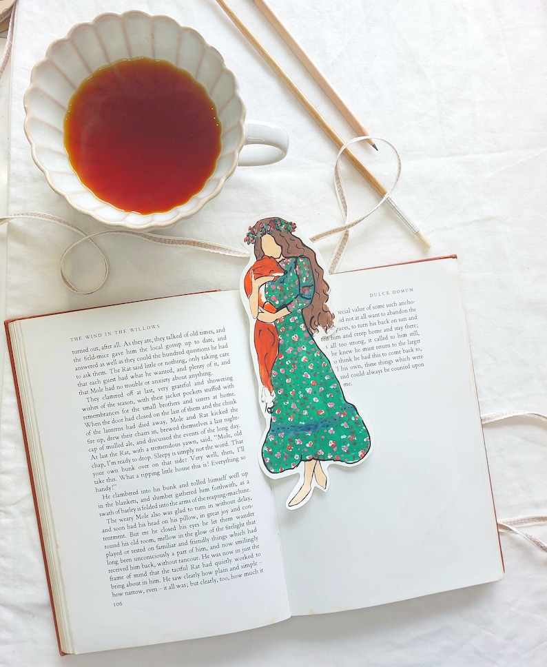 Pu&ograve; includere: Un segnalibro stravagante con una donna in un vestito floreale verde che tiene in braccio una volpe. Il segnalibro &egrave; posto su un libro aperto, insieme a una tazza di t&egrave; e elementi decorativi.