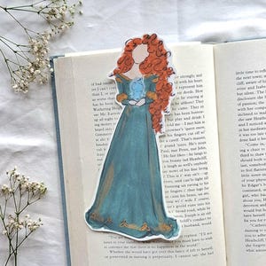 May include: A colorful bookmark featuring a cartoon illustration of a woman in a teal dress with red hair, placed on an open book. The bookmark is a whimsical accessory for readers.