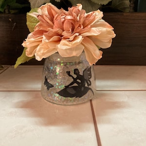 May include: A decorative glass jar with a fairy silhouette and glitter, topped with a large peach-colored artificial flower. The jar is filled with silver glitter and has a black fairy design.