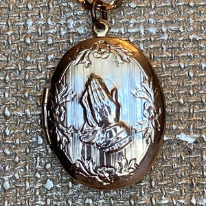 May include: Gold-toned oval locket pendant on a matching chain. The locket features an engraved design of praying hands, surrounded by floral accents. The chain is a delicate, linked style.