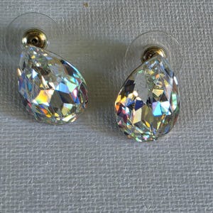 May include: A pair of clear, faceted teardrop-shaped earrings. The earrings have a gold-colored post and a clear plastic backing. The earrings are highly reflective, with a rainbow of colors visible in the facets.