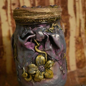 May include: A decorative jar with a textured, metallic finish in shades of purple, gray, and gold. The jar is adorned with a gold-colored floral design and a rope-wrapped lid. The overall aesthetic is ornate and handcrafted.