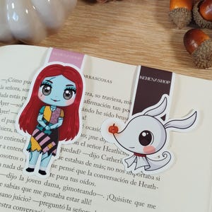 Puede incluir: Dos marcadores con temática de personajes están colocados en un libro abierto. Un marcador presenta un personaje de dibujos animados con cabello rojo y un vestido de retazos. El otro marcador representa un perro fantasma de dibujos animados. Calabazas, bellotas y una piña decoran el fondo.