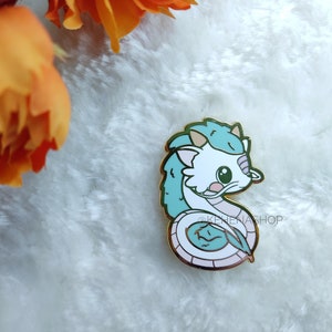 May include: A white and blue enamel pin featuring a cartoon dragon with a curled tail. The dragon has a light blue body, white head, and pink accents. The pin is on a white fuzzy surface.