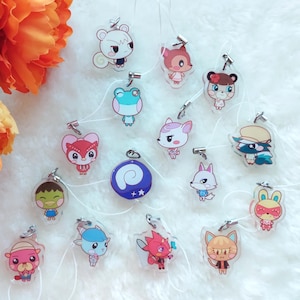 Puede incluir: Colección de colgantes acrílicos con varios personajes de animales de dibujos animados. Cada colgante transparente tiene un diseño colorido y está unido a un cierre plateado. Los colgantes están dispuestos sobre una superficie blanca y esponjosa, con flores naranjas y rojas en el fondo.