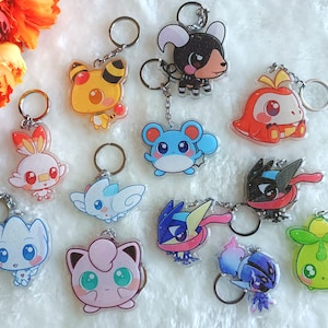May include: A collection of 11 colorful cartoon character keychains. The keychains are made of clear acrylic and feature various characters, including a pink and blue character with a flower, a yellow character with a red nose, a blue character with a red nose, a green character with a red nose, a black character with a red nose, a red character with a yellow nose, a white character with a red nose, a white character with a blue nose, a pink character with a blue nose, a blue character with a yellow nose, and a green character with a yellow nose.