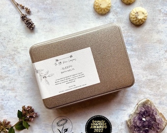 May include: A gold tin with a white label that reads "The Free Grace Company - SLEEPY - BATH MELTS" and a description of the bath melts. The tin is sitting on a white surface with three white bath melts and a purple amethyst crystal. The image also includes a vegan logo and a beauty shortlist award logo.