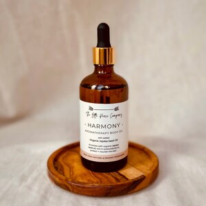 May include: A brown glass bottle with a gold lid and dropper, labeled "The Peace Company HARMONY AROMATHERAPY BODY OIL with added Organic Jojoba Seed Oil". The bottle is sitting on a wooden coaster.
