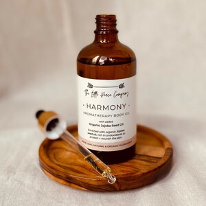 May include: A brown glass bottle with a dropper, labeled "The Little Peace Company. HARMONY. AROMATHERAPY BODY OIL with added Organic Jojoba Seed Oil. Enriched with organic Jojoba seed oil, rich in antioxidants to protect + nourish the skin. MADE USING NATURAL & ORGANIC INGREDIENTS."