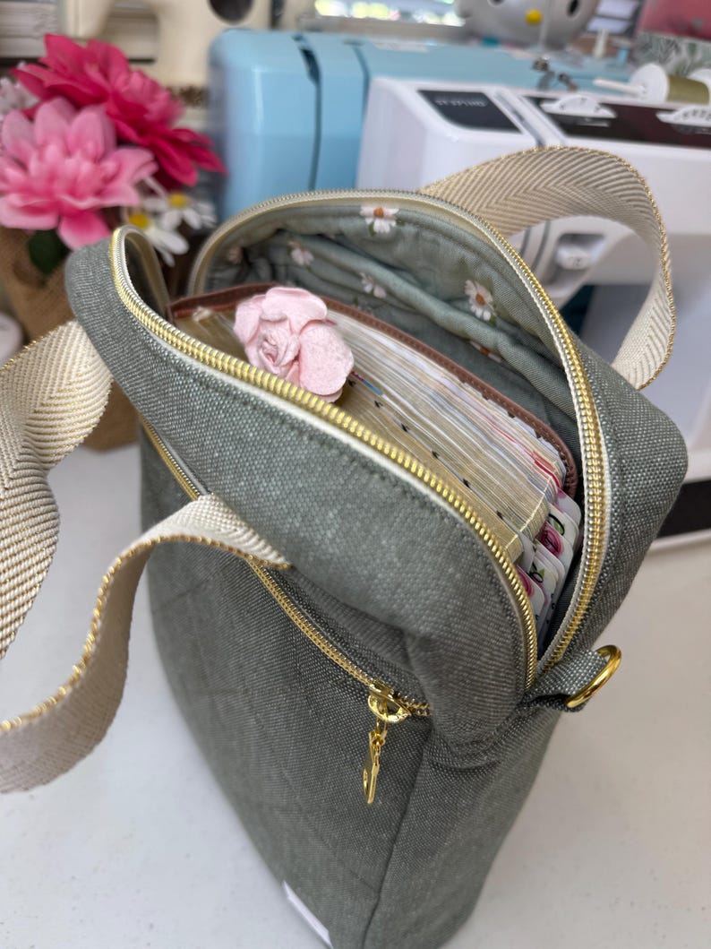 Puede incluir: Una bolsa de lona verde con una cremallera dorada y una cadena dorada. La bolsa est&aacute; abierta y llena de una pila de papeles y una flor rosa.