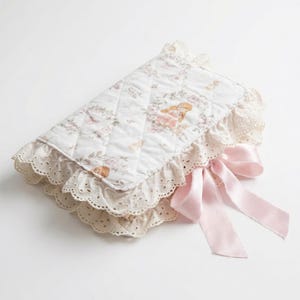 May include: A quilted, white baby changing pad with a floral pattern and a decorative lace trim. The pad features a pink satin bow and a design with a baby angel. The pad is designed for baby care.