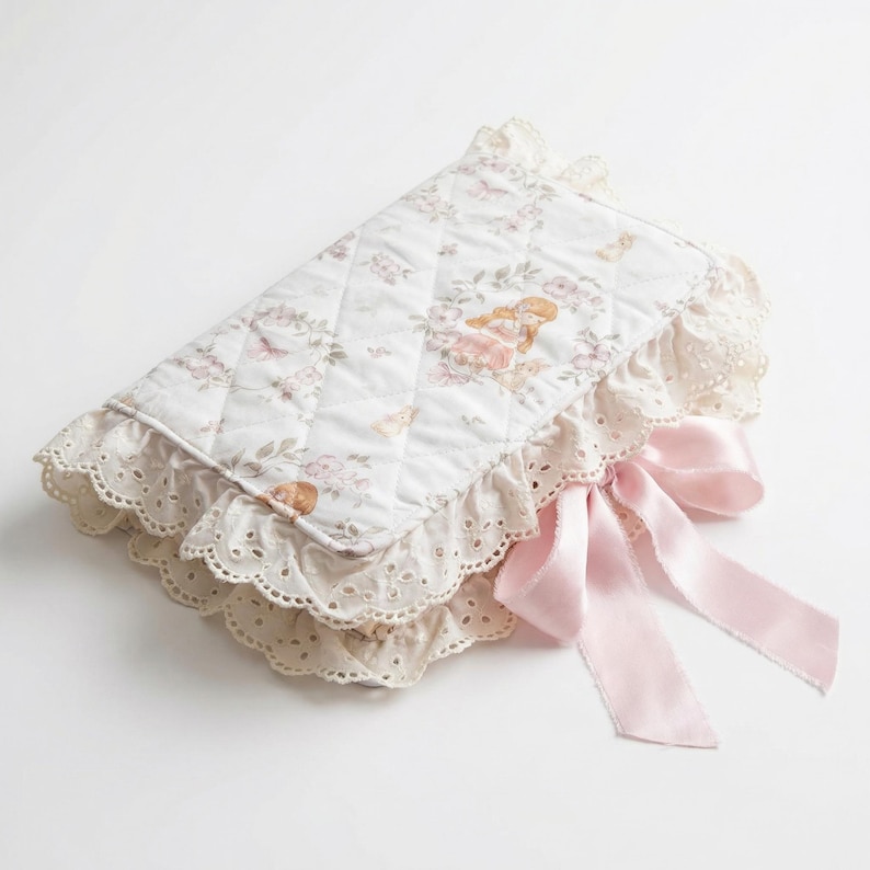 May include: A quilted, white baby changing pad with a floral pattern and a decorative lace trim. The pad features a pink satin bow and a design with a baby angel. The pad is designed for baby care.