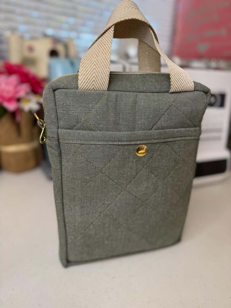 Puede incluir: Una bolsa de lona verde acolchada con cierre de cremallera y asa superior. La bolsa tiene un peque&ntilde;o bolsillo en la parte delantera y un bot&oacute;n dorado.