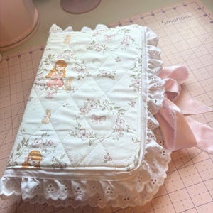May include: A quilted, floral-patterned journal with a white eyelet lace trim and a pink satin ribbon closure. The cover features a repeating pattern of girls, bunnies, and flowers in soft pastel colors. The journal is closed.