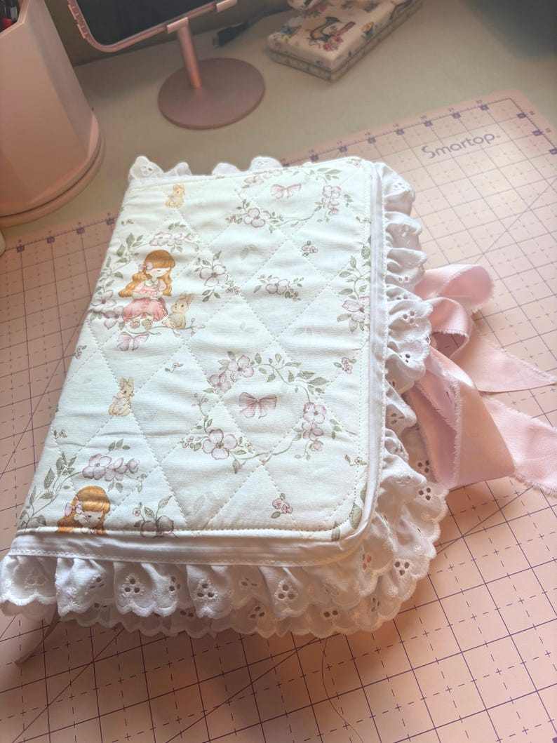 May include: A quilted, floral-patterned journal with a white eyelet lace trim and a pink satin ribbon closure. The cover features a repeating pattern of girls, bunnies, and flowers in soft pastel colors. The journal is closed.