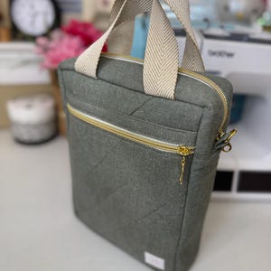 Puede incluir: Una bolsa de lona gris con cierre de cremallera y asa de color crema. La bolsa tiene un dise&ntilde;o acolchado y un tirador de cremallera dorado.