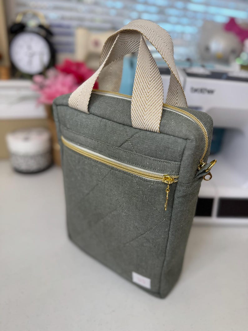 Puede incluir: Una bolsa de lona gris con cierre de cremallera y asa de color crema. La bolsa tiene un dise&ntilde;o acolchado y un tirador de cremallera dorado.