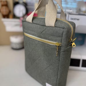 Puede incluir: Una funda para port&aacute;til acolchada gris con cierre de cremallera y asa de color crema. La funda tiene una cremallera dorada y un tirador dorado.