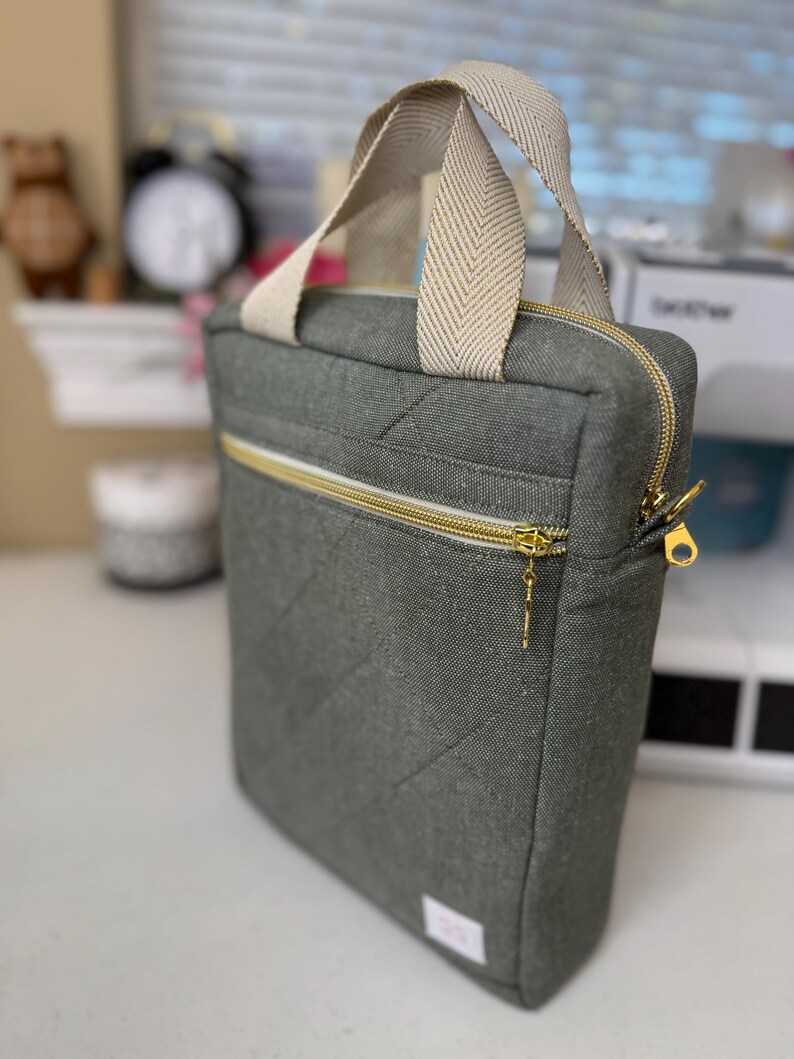 Puede incluir: Una funda para port&aacute;til acolchada gris con cierre de cremallera y asa de color crema. La funda tiene una cremallera dorada y un tirador dorado.