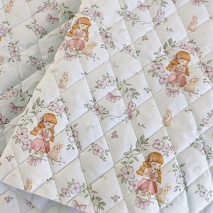 May include: White quilted fabric with a repeating pattern of a girl in a pink dress with a bunny, flowers, and bows. The fabric is stitched in a diamond pattern, creating a textured surface. The overall design has a soft, vintage aesthetic.