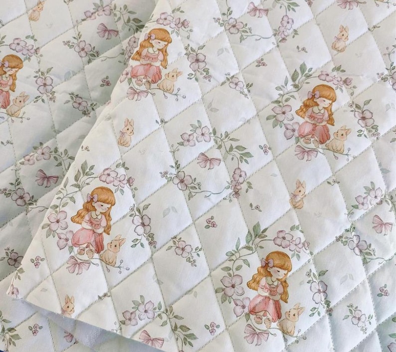 May include: White quilted fabric with a repeating pattern of a girl in a pink dress with a bunny, flowers, and bows. The fabric is stitched in a diamond pattern, creating a textured surface. The overall design has a soft, vintage aesthetic.