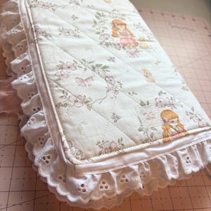May include: A quilted, off-white journal cover with a floral and girl illustration pattern. The cover is trimmed with white eyelet lace and a satin ribbon. The journal is a soft, feminine accessory.